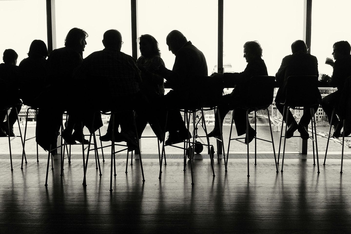 Gruppe im Gegenlicht an einem langen Tisch vor einer Fensterfront, nur als Silhouetten erkennbar