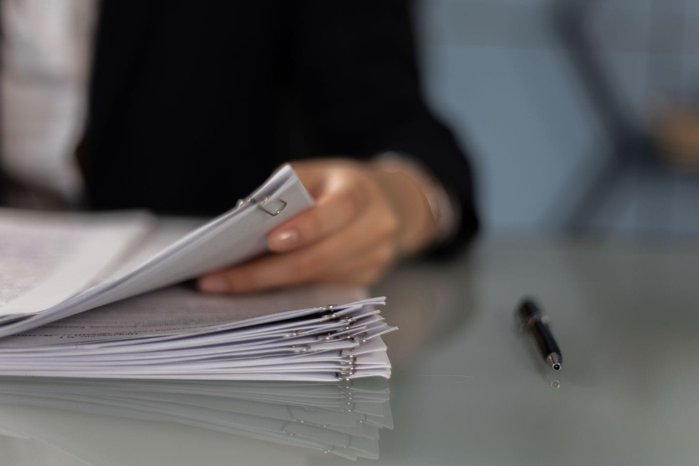 Hand blättert in einem Stapel geklammerter Papierbögen, daneben ein Kugelschreiber