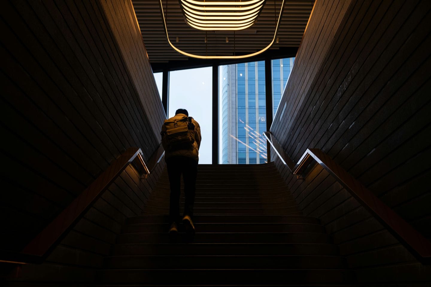 Person mit Rucksack steigt eine dunkle Treppe zu einer Glasfront hinauf, dahinter ein Hochhaus